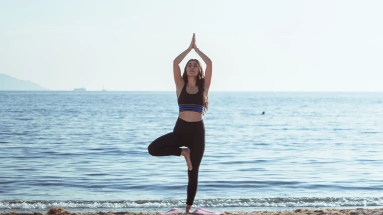 Woman doing yoga tree pose on a beach with calm ocean water in the background.