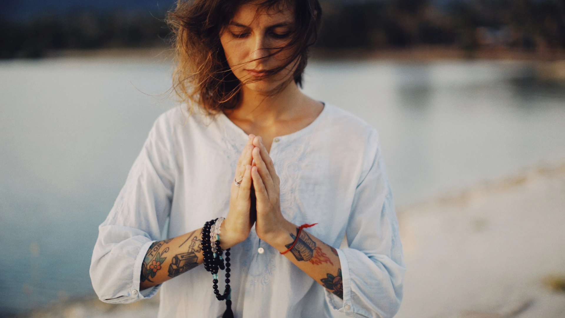 Woman with closed eyes and hands in prayer position, wearing mala beads, standing by a lake at dusk